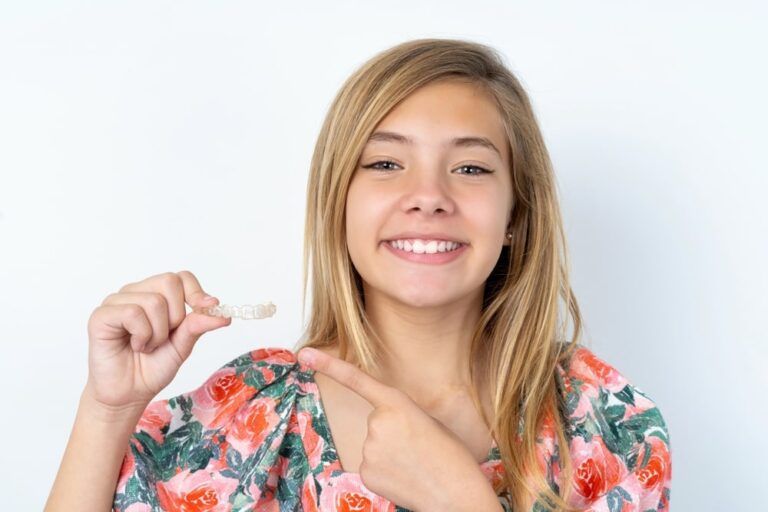 beautiful caucasian teen girl wearing flowered blouse over white wall holding an invisible aligner and pointing at it