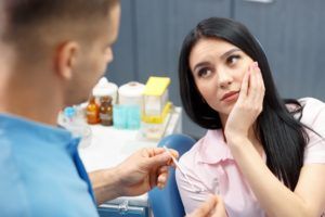 Woman sitting in dental chair talking to dentist and holding her cheek in pain