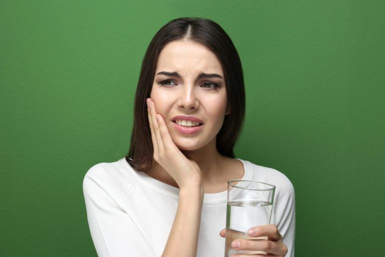 Brunette woman on green background holding a glass of water and one hand and her cheek in the other with a pained expression
